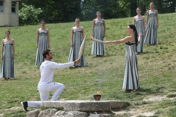 扮演最高女祭司的演員瑪麗·米娜（前右）點(diǎn)燃第一棒火炬手——希臘運(yùn)動員、東京奧運(yùn)會男子賽艇項(xiàng)目金牌得主斯特凡諾斯·恩圖斯科斯手中的火炬。新華社記者 趙丁喆 攝